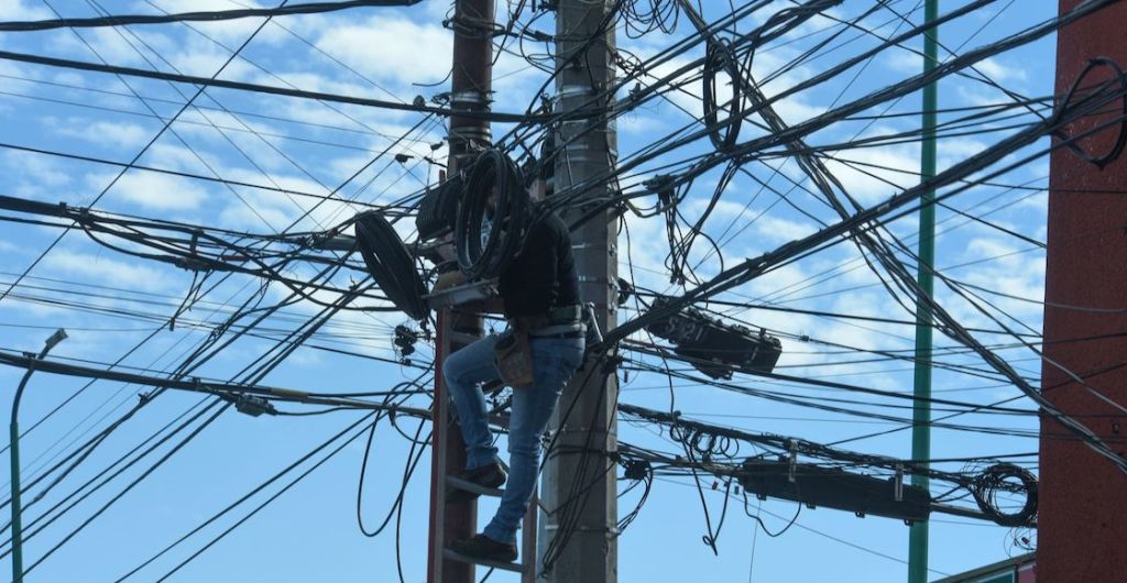 Un hombre trabaja en lo alto de un poste entre la telaraña de cables.