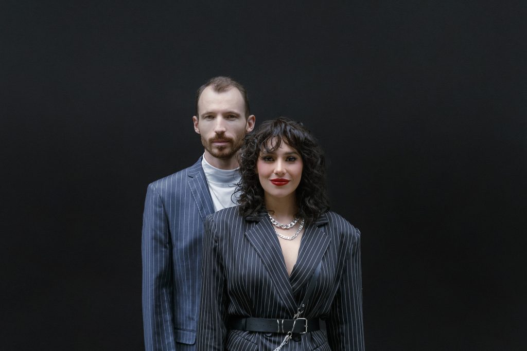 Marissa Mur posando junto al músico Joséan Log con trajes a rayas sobre fondo oscuro.
