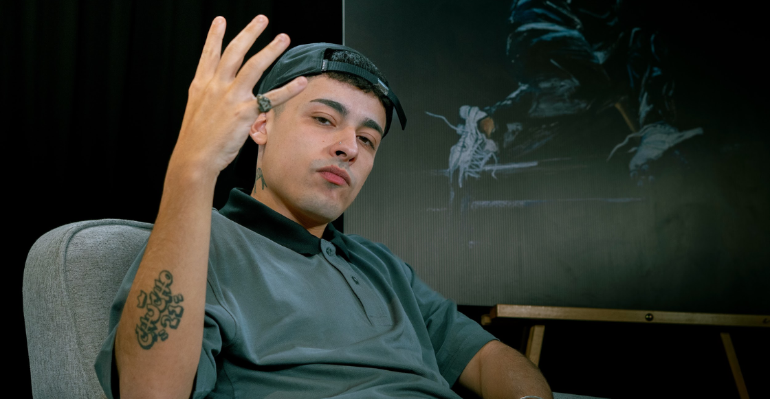 Trueno sentado en una silla durante una entrevista, haciendo una seña con la mano y usando gorra negra y playera gris.