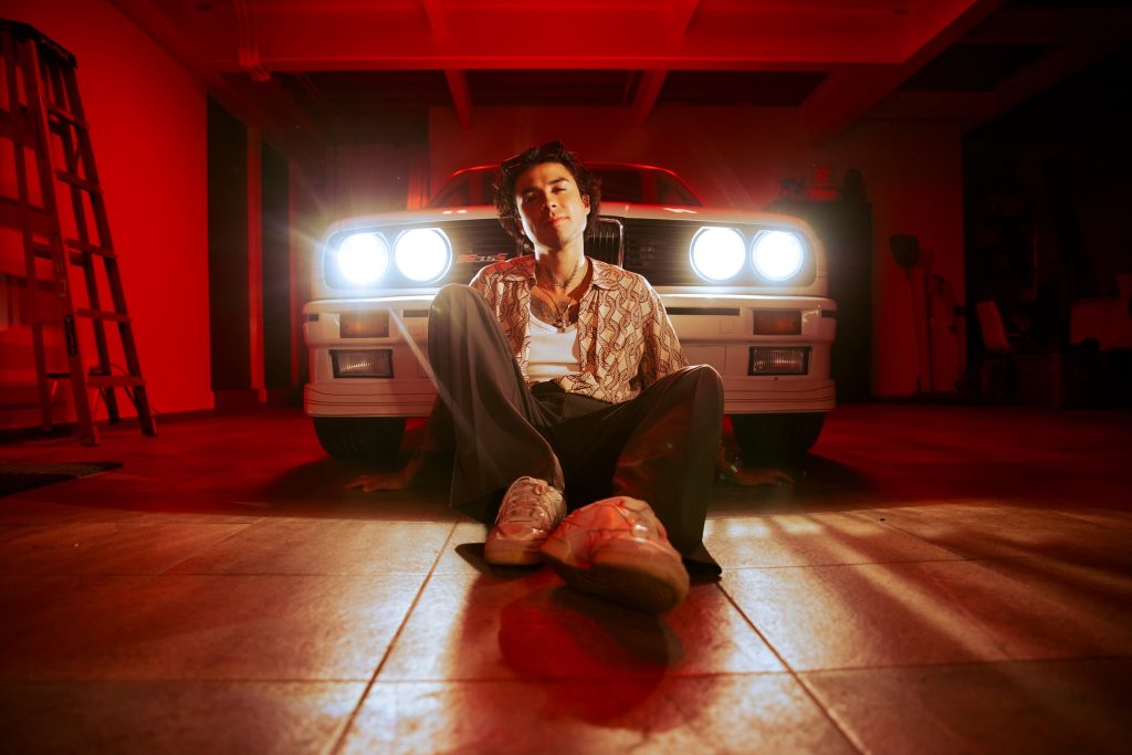 Mario Bautista posa sentado frente a un auto clásico blanco con luces encendidas en una sesión fotográfica con iluminación roja.