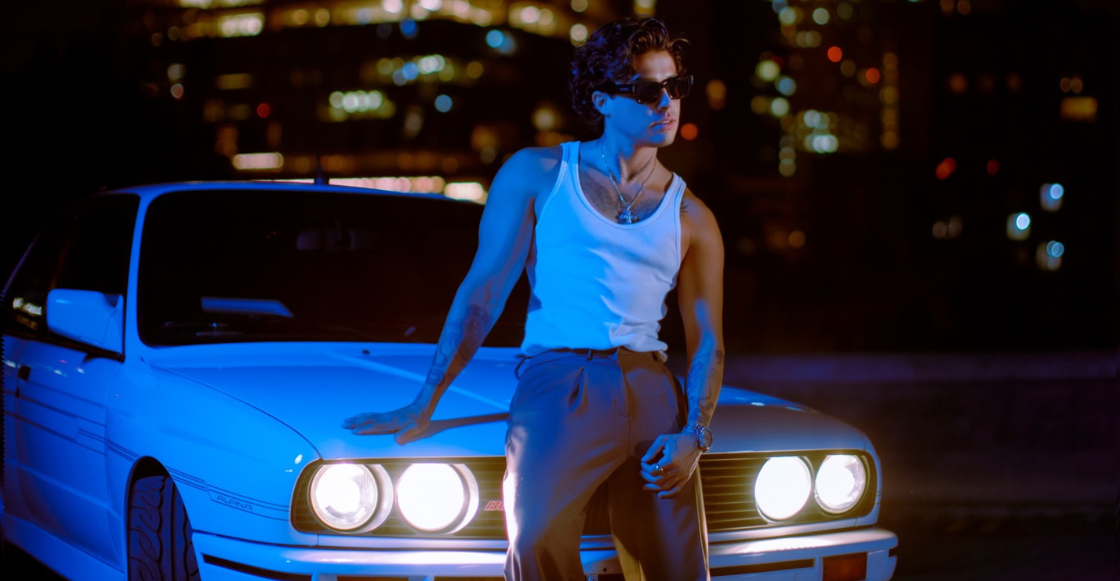 Mario Bautista posa junto a un auto blanco clásico durante una sesión nocturna con luces azules y fondo urbano desenfocado.