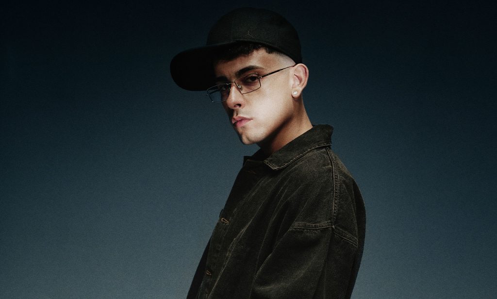 Retrato de Trueno con gorra negra ladeada, lentes delgados y chamarra oscura sobre un fondo azul oscuro.