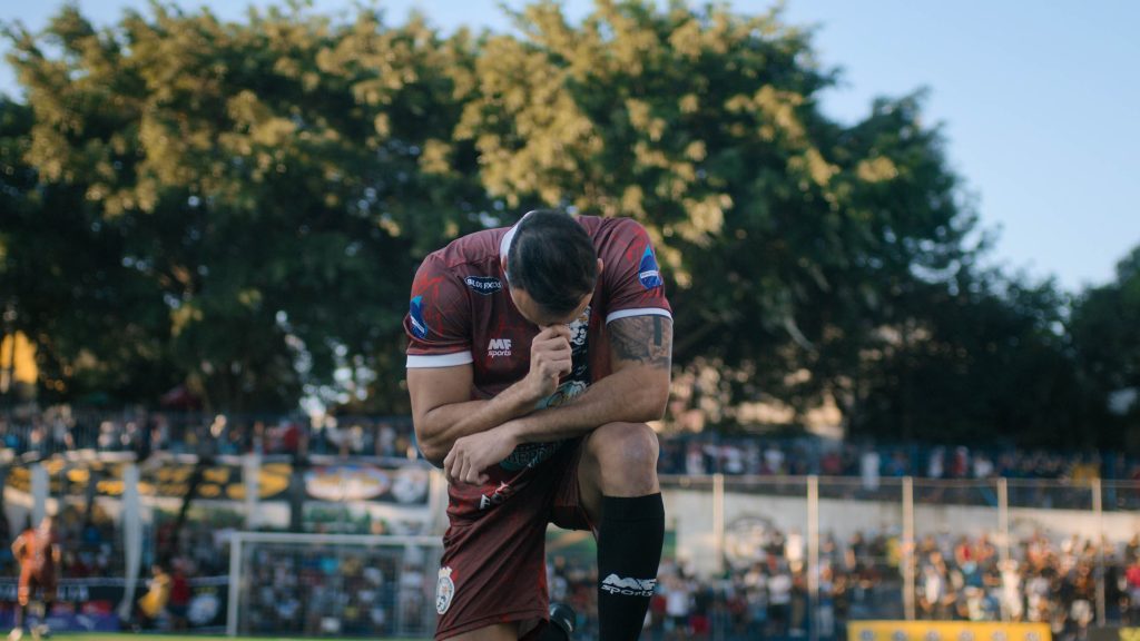 Jugador de futbol amateur se arrodilla en una cancha durante una escena de Várzea: The Root of the Game de Netflix.