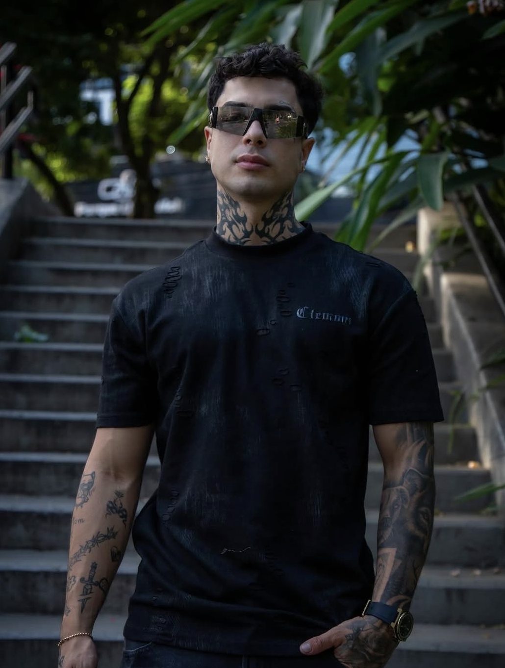 Naim Darrechi posando al aire libre con playera negra, lentes de sol y tatuajes visibles.