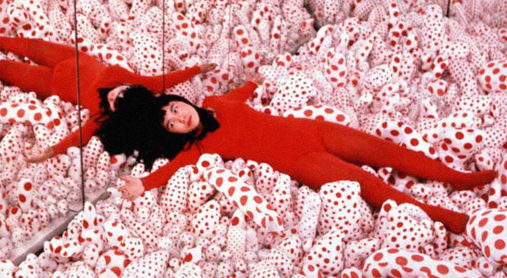Yayoi Kusama recostada con un traje rojo sobre esculturas blandas blancas con lunares rojos en su instalación Infinity Mirror Room Phalli's Field.