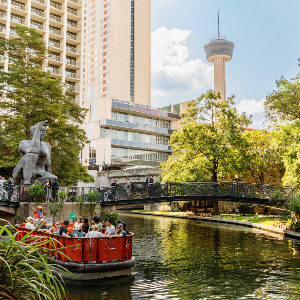 Barcaza de turistas navegando por el River Walk con la Tower of the Americas al fondo, en San Antonio, Texas.