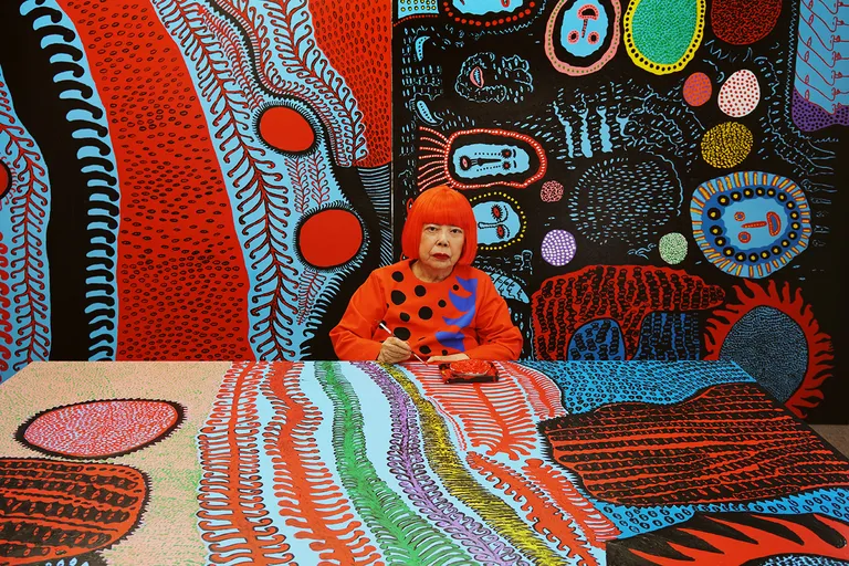 Yayoi Kusama sentada en su estudio trabajando con un pincel sobre un lienzo colorido con sus característicos patrones y lunares, rodeada de sus obras vibrantes.
