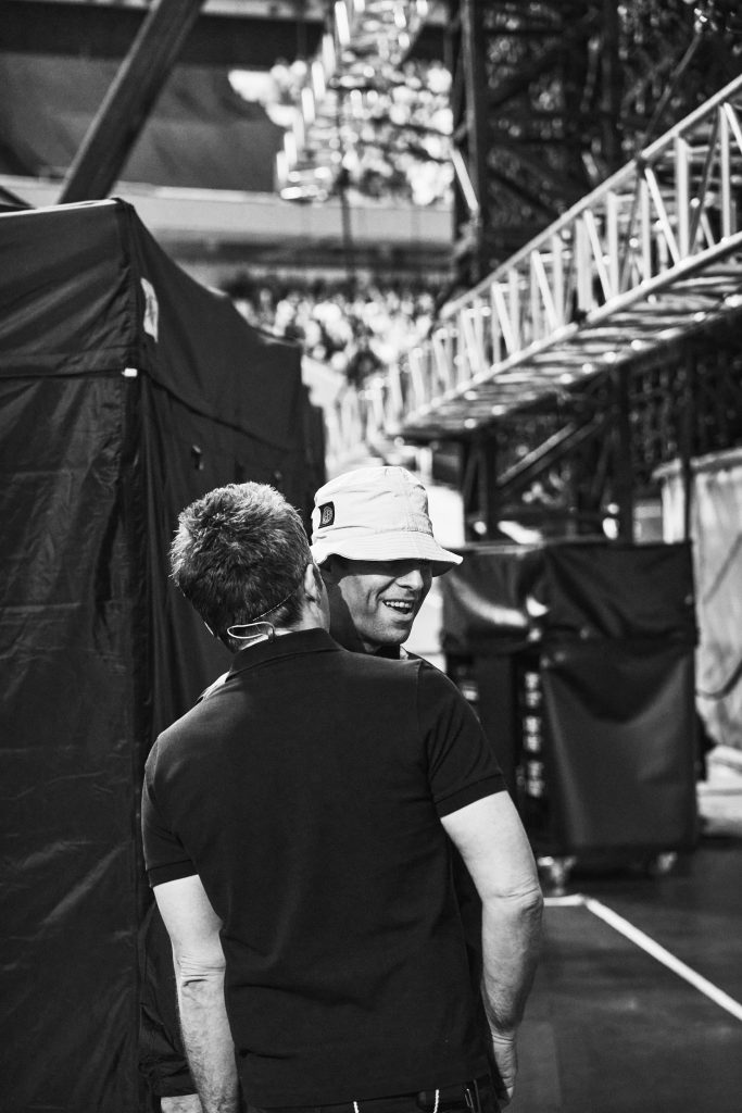 Fotografía en blanco y negro de Liam Gallagher sonriendo y usando un sombrero de pescador, frente a Noel Gallagher que se encuentra de espaldas, en una zona de backstage.