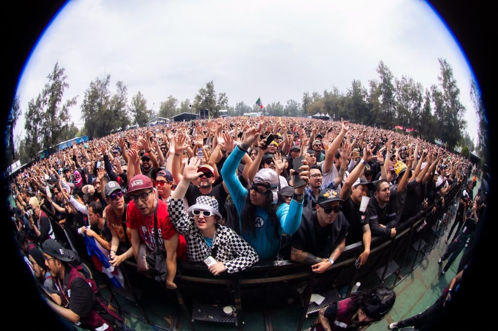 Una toma panorámica de gran angular que muestra una inmensa multitud de fans de música en el Vans Warped Tour México, muchos haciendo señas con las manos (como el símbolo de 3OH!3 o el 'rock on'), bajo un cielo parcialmente nublado. La imagen captura la energía y la escala del festival.