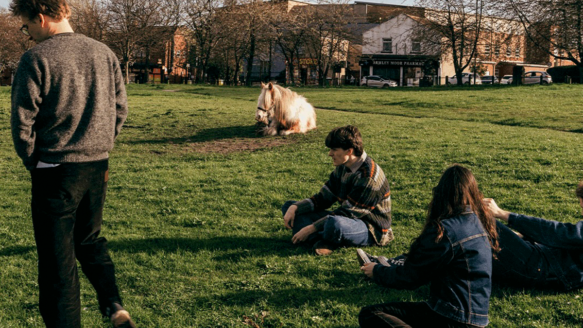 Integrantes de la banda Westside Cowboy relajándose en un parque verde; tres de ellos sentados en el pasto y uno de pie, con un caballo claro al fondo.