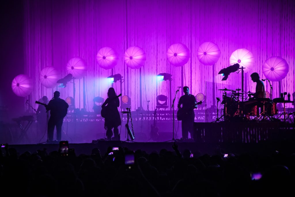 Siluetas de Of Monsters and Men sobre el escenario del Pepsi Center con iluminación violeta y esferas luminosas de escenografía al fondo.