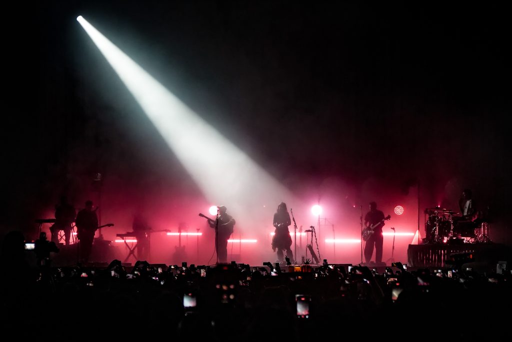 Texto alternativo:
Siluetas de Of Monsters and Men iluminadas por un reflector blanco cenital en el escenario del Pepsi Center con luces rojas al fondo.